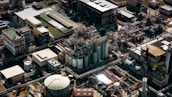 An aerial view of a large industrial complex with numerous buildings, pipelines, and storage tanks. The structures are closely packed, showcasing a network of interconnected pipes and machinery. The scene is densely composed with various rectangular rooftops and cylindrical silos.