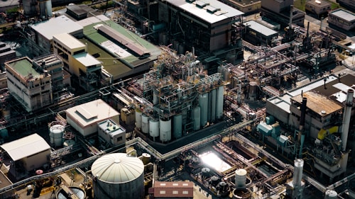 An aerial view of a large industrial complex with numerous buildings, pipelines, and storage tanks. The structures are closely packed, showcasing a network of interconnected pipes and machinery. The scene is densely composed with various rectangular rooftops and cylindrical silos.