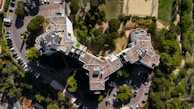 An aerial view captures a large, uniquely shaped residential or commercial building surrounded by a number of trees and a parking lot. The structure features irregular angles and multiple levels with rooftop access. Several parked cars are visible in the parking area, and nearby, a sports field is outlined by a metal fence.