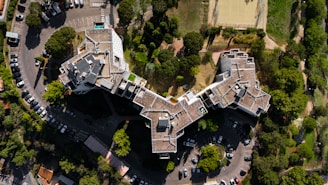 An aerial view captures a large, uniquely shaped residential or commercial building surrounded by a number of trees and a parking lot. The structure features irregular angles and multiple levels with rooftop access. Several parked cars are visible in the parking area, and nearby, a sports field is outlined by a metal fence.