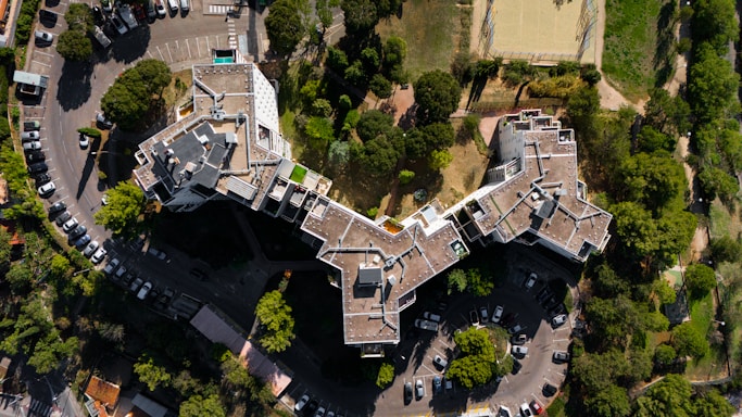 An aerial view captures a large, uniquely shaped residential or commercial building surrounded by a number of trees and a parking lot. The structure features irregular angles and multiple levels with rooftop access. Several parked cars are visible in the parking area, and nearby, a sports field is outlined by a metal fence.