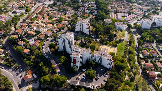 An aerial view of a residential area featuring several mid-rise apartment buildings surrounded by lush greenery. The image also includes a neighborhood with rows of smaller houses, streets, and cars parked along the roads. The landscape is dotted with trees and open green spaces, indicating a suburban setting.