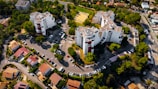 Several modern apartment buildings are surrounded by greenery and a well-planned road network. The area includes a series of parked cars, individual houses with red roofs, and a sports field in the middle of the complex. Trees and shrubs are abundant, contributing to a lush environment.