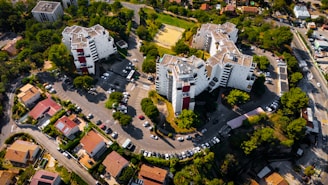 Several modern apartment buildings are surrounded by greenery and a well-planned road network. The area includes a series of parked cars, individual houses with red roofs, and a sports field in the middle of the complex. Trees and shrubs are abundant, contributing to a lush environment.