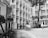 A group of students in uniform are walking through a school courtyard surrounded by multi-story buildings. The scene includes a tree and a fence adorned with potted plants. The image is in black and white, giving it a classic and timeless look.