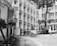 A group of students in uniform are walking through a school courtyard surrounded by multi-story buildings. The scene includes a tree and a fence adorned with potted plants. The image is in black and white, giving it a classic and timeless look.