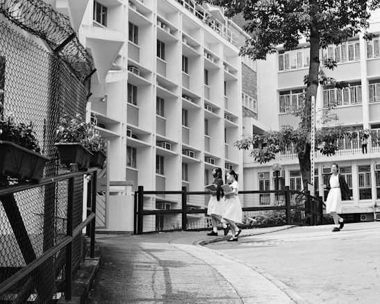 A group of students in uniform are walking through a school courtyard surrounded by multi-story buildings. The scene includes a tree and a fence adorned with potted plants. The image is in black and white, giving it a classic and timeless look.