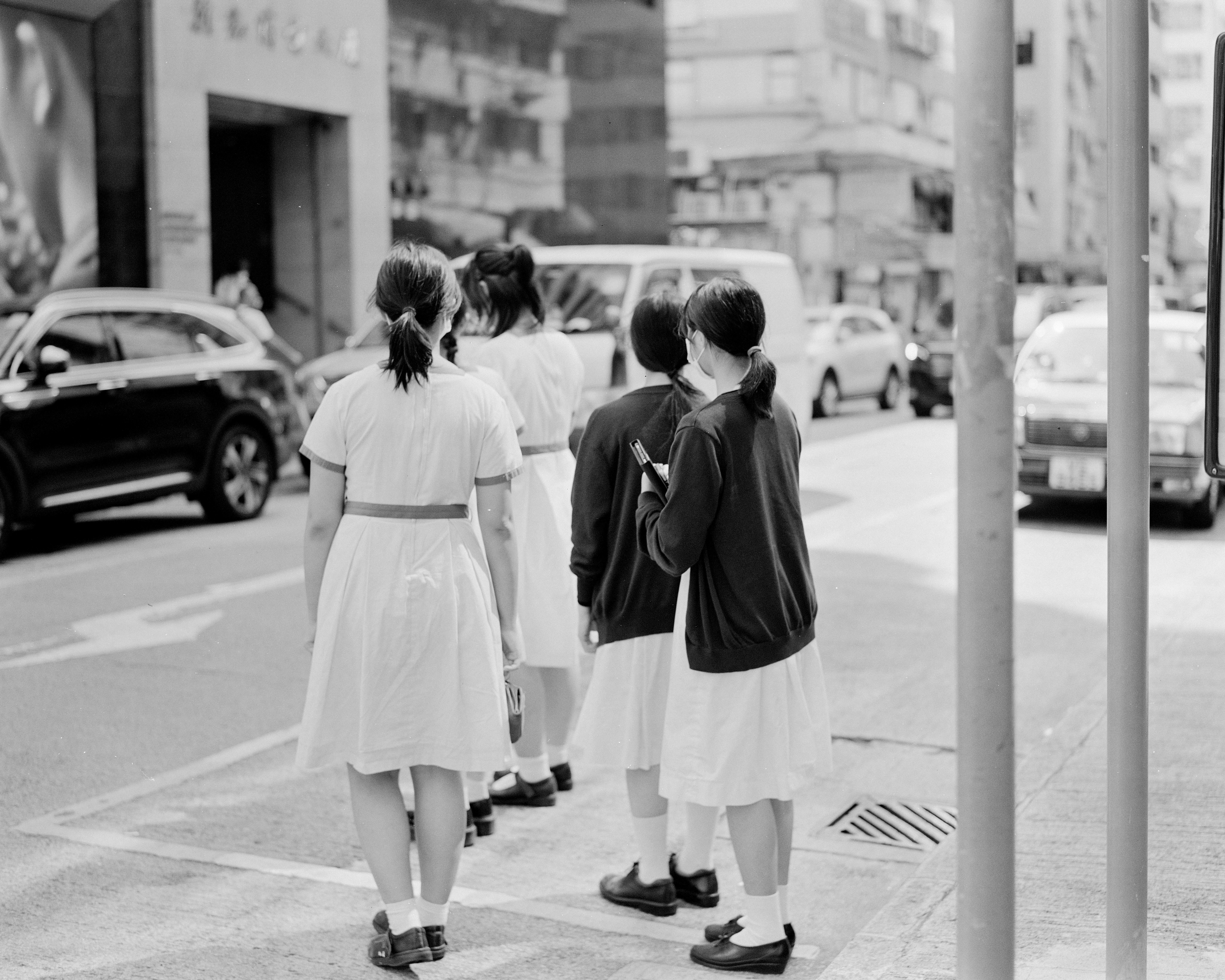 A group of girls in uniform dresses are standing at the side of a city street. They are facing away from the camera and appear to be waiting to cross the road. The surroundings include various vehicles such as cars and a van, as well as urban buildings and traffic signs.