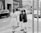 A group of girls in uniform dresses are standing at the side of a city street. They are facing away from the camera and appear to be waiting to cross the road. The surroundings include various vehicles such as cars and a van, as well as urban buildings and traffic signs.