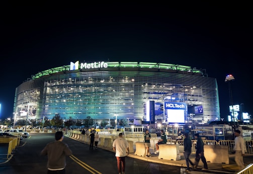 A large, illuminated stadium at night with bright signage including the MetLife and HCLTech logos. People are walking towards the entrance, and the surroundings are well-lit, with barriers guiding pedestrian traffic.
