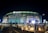A large, illuminated stadium at night with bright signage including the MetLife and HCLTech logos. People are walking towards the entrance, and the surroundings are well-lit, with barriers guiding pedestrian traffic.