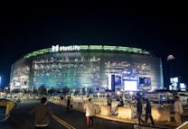 A large, illuminated stadium at night with bright signage including the MetLife and HCLTech logos. People are walking towards the entrance, and the surroundings are well-lit, with barriers guiding pedestrian traffic.
