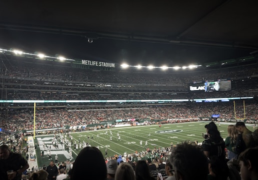 A large stadium with a football field is filled with a crowd of spectators. The field is set for an American football game with players and officials on the field. Bright stadium lights illuminate the area, and a large sign reads 'MetLife Stadium'. Fans are seated in the stands, some standing and watching the game.