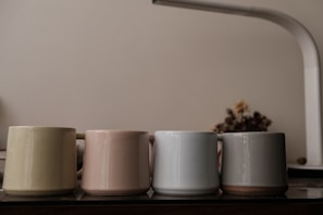 Four ceramic mugs of different muted colors are lined up on a wooden surface. A soft yet bright light illuminates the scene, creating gentle reflections on the smooth surfaces of the mugs. In the background, there is a blurred figure of dried flowers and a white, arching lamp creating an atmosphere of calm sophistication.