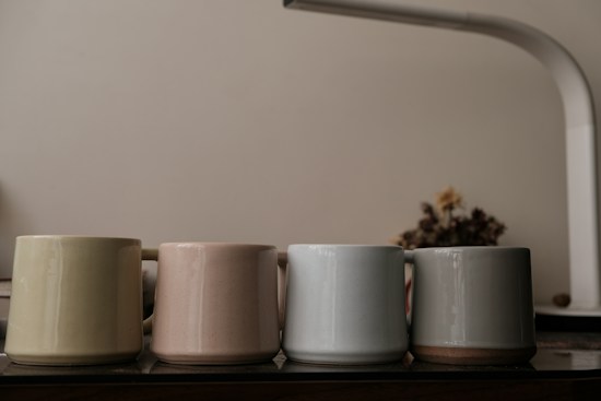 Four ceramic mugs of different muted colors are lined up on a wooden surface. A soft yet bright light illuminates the scene, creating gentle reflections on the smooth surfaces of the mugs. In the background, there is a blurred figure of dried flowers and a white, arching lamp creating an atmosphere of calm sophistication.