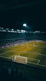 A nighttime soccer match is in progress in a large, well-lit stadium filled with spectators. Players are scattered across the field, with a goalkeeper standing near the goal post. The atmosphere appears vibrant with a crowd watching the game intensely.