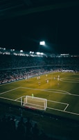 A nighttime soccer match is in progress in a large, well-lit stadium filled with spectators. Players are scattered across the field, with a goalkeeper standing near the goal post. The atmosphere appears vibrant with a crowd watching the game intensely.