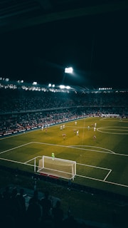A nighttime soccer match is in progress in a large, well-lit stadium filled with spectators. Players are scattered across the field, with a goalkeeper standing near the goal post. The atmosphere appears vibrant with a crowd watching the game intensely.