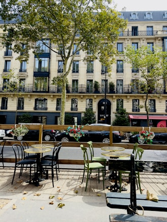 Outdoor cafe seating area with several tables and chairs, situated on a wooden deck. The setting is adjacent to a street with parked cars. Behind the street is a multi-story building with elegant balconies and large windows. Trees with green foliage partially obscure the view.