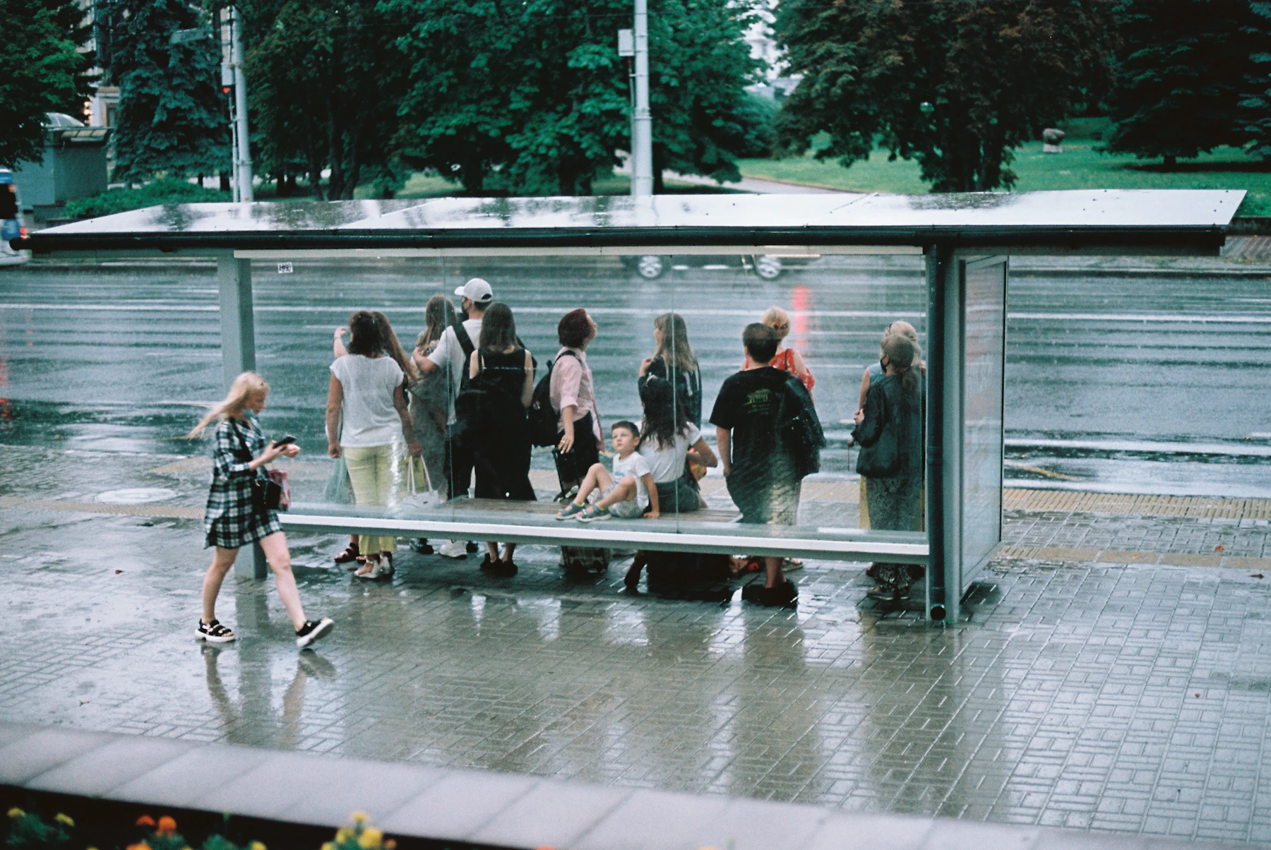 A group of riders standing under a glass bus stop canopy on a wet day, the pavement shining with rain