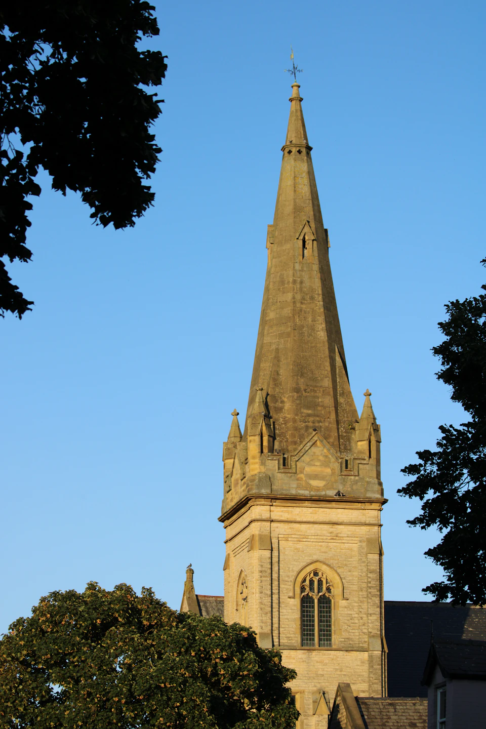 The image depicts a tall, light brown stone church with a pointed steeple and a cross at its pinnacle. The church is situated amidst lush green trees, creating a serene and tranquil atmosphere. The perspective of the photo is from below, looking up towards the church, emphasizing its height and grandeur against the backdrop of the sky.