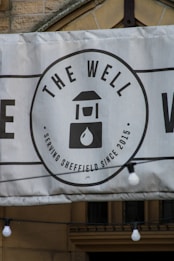 A banner displaying a logo with the text 'THE WELL' and an image of a water well with a droplet. The banner indicates the establishment has been serving Sheffield since 2015. It is mounted on a stone building and features decorative light bulbs hanging in front of it.