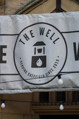 A banner displaying a logo with the text 'THE WELL' and an image of a water well with a droplet. The banner indicates the establishment has been serving Sheffield since 2015. It is mounted on a stone building and features decorative light bulbs hanging in front of it.