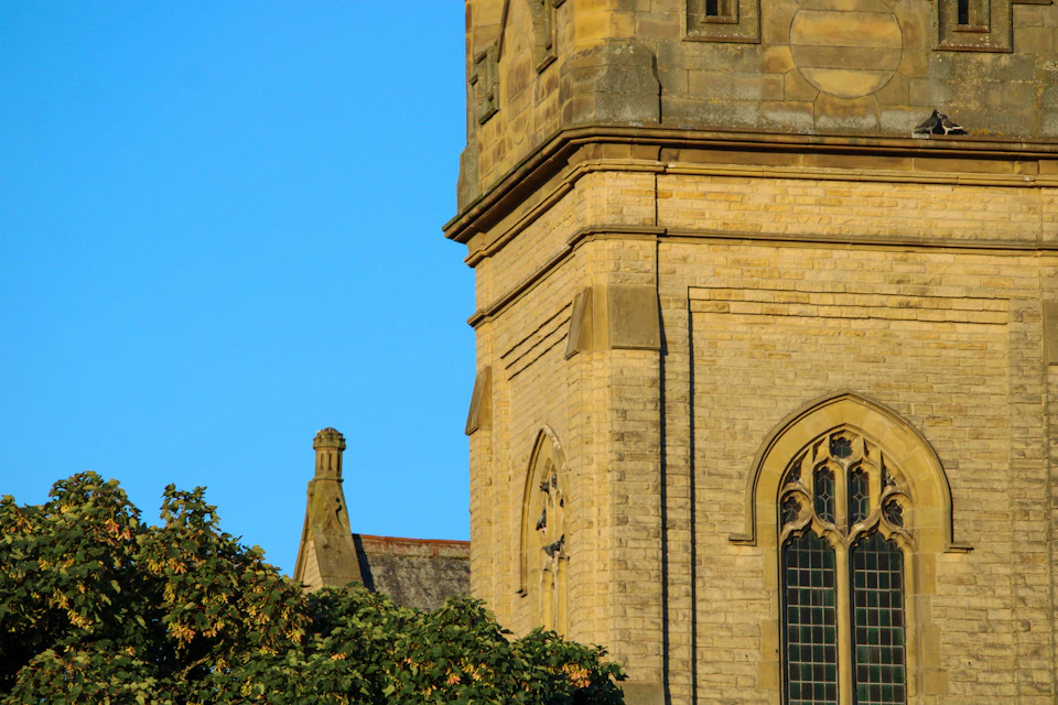The image depicts a stone church with a tall tower and a large arched window, set against a clear blue sky. The church's architecture features multiple levels, including a smaller tower on top. In front of the church, there are trees that add a touch of nature to the scene.