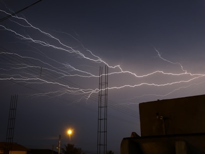 An intense lightning storm is captured with multiple bright, jagged lightning bolts illuminating the night sky. Electrical wires and streetlights are faintly visible in the foreground, contrasting with the dark silhouettes of buildings.