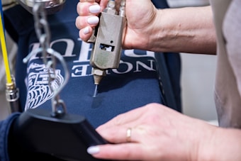 A person is pressing a metallic tool onto a dark blue cloth, possibly involved in printing or embroidery. The cloth features white text and a graphic design. The person’s hands are visible, and they have white nail polish.