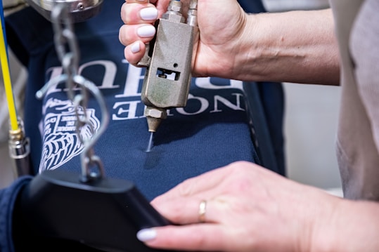 A person is pressing a metallic tool onto a dark blue cloth, possibly involved in printing or embroidery. The cloth features white text and a graphic design. The person’s hands are visible, and they have white nail polish.
