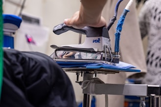 A close-up view of a handheld industrial iron in use, with a person's hand gripping it firmly. The iron is placed on a padded, blue surface. The background contains blurred elements indicating a garment and some other equipment.