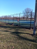 A person is playing tennis on an outdoor court surrounded by a tall chain-link fence. The court has a green surface, and there is a tennis bag on the ground. The surrounding area features bare trees and a clear blue sky, indicating a cold season.