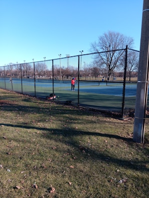 A person is playing tennis on an outdoor court surrounded by a tall chain-link fence. The court has a green surface, and there is a tennis bag on the ground. The surrounding area features bare trees and a clear blue sky, indicating a cold season.