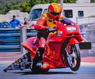 A person wearing a yellow and orange racing suit and helmet is riding a red racing motorcycle. The motorcycle has numerous stickers and decals on it, creating a dynamic and intense scene. In the background, there are blurred figures and vehicles, indicating motion and speed.
