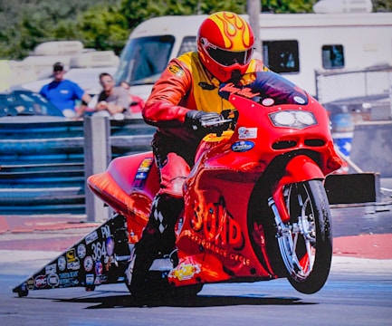 A person wearing a yellow and orange racing suit and helmet is riding a red racing motorcycle. The motorcycle has numerous stickers and decals on it, creating a dynamic and intense scene. In the background, there are blurred figures and vehicles, indicating motion and speed.