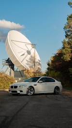 A white sedan is parked on a paved area surrounded by greenery. In the background, a large satellite dish is prominently positioned, capturing attention with its size and structure. The sky is clear with a few clouds scattered, and the trees are in autumn colors, adding a mix of green and orange hues to the scene.