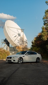 A white sedan is parked on a paved area surrounded by greenery. In the background, a large satellite dish is prominently positioned, capturing attention with its size and structure. The sky is clear with a few clouds scattered, and the trees are in autumn colors, adding a mix of green and orange hues to the scene.