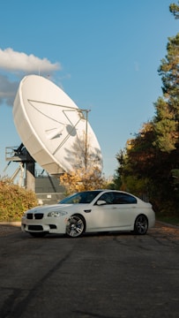 A white sedan is parked on a paved area surrounded by greenery. In the background, a large satellite dish is prominently positioned, capturing attention with its size and structure. The sky is clear with a few clouds scattered, and the trees are in autumn colors, adding a mix of green and orange hues to the scene.