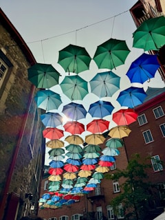 A collection of colorful umbrellas is suspended in the air, creating a vibrant canopy effect in an urban setting. These umbrellas, in shades of green, blue, red, and beige, hang between buildings made of brick and stone. Sunlight peeks through, casting decorative shadows.