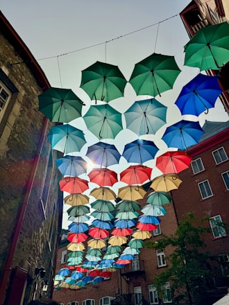 A collection of colorful umbrellas is suspended in the air, creating a vibrant canopy effect in an urban setting. These umbrellas, in shades of green, blue, red, and beige, hang between buildings made of brick and stone. Sunlight peeks through, casting decorative shadows.