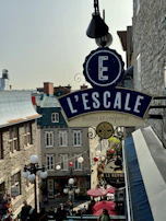 An outdoor scene in a European-style street featuring a large sign for 'L'ESCALE bistro et crêperie' hanging prominently. The street is lined with stone buildings with classic architectural details, and there are several lamps and decorations, such as hanging lights and umbrellas. People are visible walking and dining, creating a lively atmosphere.