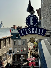 An outdoor scene in a European-style street featuring a large sign for 'L'ESCALE bistro et cr&ecirc;perie' hanging prominently. The street is lined with stone buildings with classic architectural details, and there are several lamps and decorations, such as hanging lights and umbrellas. People are visible walking and dining, creating a lively atmosphere.