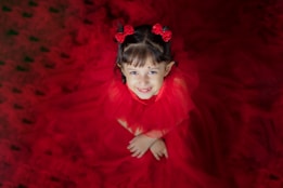 A young girl smiles warmly while dressed in an elaborate red gown with layers of fabric surrounding her. Her hair is styled with red floral accessories, complementing her outfit. She is looking upwards towards the camera, creating a charming and joyful atmosphere.