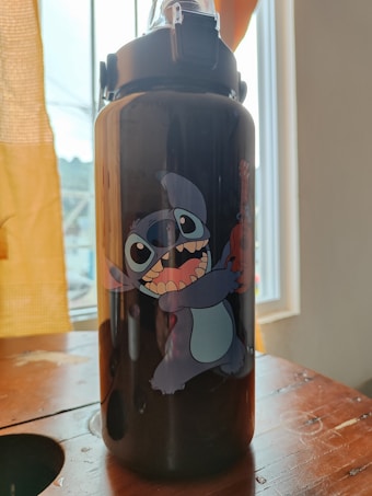 A dark-colored water bottle with a cheerful cartoon character printed on it, placed on a wooden surface near a window with light curtains.