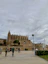 A group of people ride bicycles along a paved path in front of a large Gothic-style cathedral. The sky is overcast, and a fountain is visible on the right side. Palm trees and a historic stone wall are also present, adding to the scenic setting.