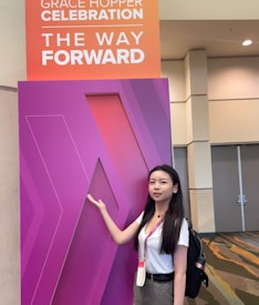 A woman stands next to a large purple sign with geometric patterns. The sign has a smaller panel above it that reads 'Grace Hopper Celebration The Way Forward'. She is wearing a white shirt, with a name tag around her neck, and has a backpack.