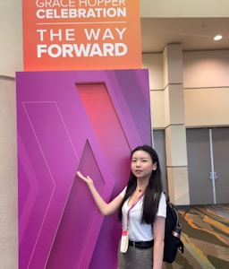 A woman stands next to a large purple sign with geometric patterns. The sign has a smaller panel above it that reads 'Grace Hopper Celebration The Way Forward'. She is wearing a white shirt, with a name tag around her neck, and has a backpack.