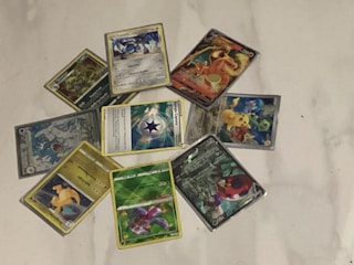 A collection of Pokémon trading cards is arranged in a circular pattern on a white surface. The cards feature various Pokémon characters, each with different illustrations and designs. Some cards have a metallic or holographic finish, adding a reflective quality to the images.