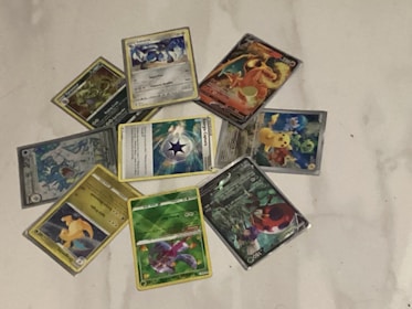 A collection of Pokémon trading cards is arranged in a circular pattern on a white surface. The cards feature various Pokémon characters, each with different illustrations and designs. Some cards have a metallic or holographic finish, adding a reflective quality to the images.